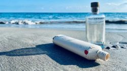 A reusable glass bottle upright on the beach. Next to it lies a plastic bottle that is damaged in some places. 