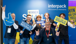 Several people holding interpack signs stand in front of a photo wall and pose together for a picture.